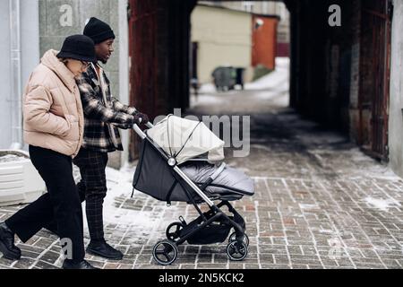 Felice famiglia interracial camminare sulla strada e spingere carrozza bambino. Concetto di famiglia interrazziale e unità tra diverse razze umane. Foto Stock