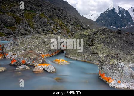 Acque calme vicino a pietre con muschio arancione contro montagne con ghiacciai e neve in Altai, Siberia. Foto Stock