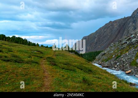 Il percorso in erba verde si estende accanto ad un torrente d'alta quota fino ad un arcobaleno nelle nuvole di tuono sulle montagne rocciose di Altai in estate. Foto Stock