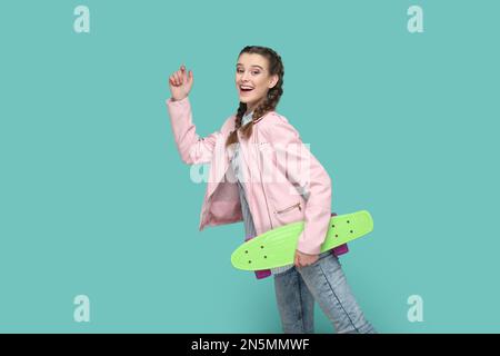 Ritratto di ragazza adolescente estremamente felice divertente con trecce indossare giacca rosa in piedi con braccio sollevato, tenendo lo skateboard in mano. Studio in interni isolato su sfondo verde. Foto Stock