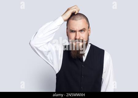 Ritratto di un bell'uomo pensieroso e penoso con barba e baffi profondi nei pensieri, tenendo la mano sulla testa, meditando. Studio in interni isolato su sfondo grigio. Foto Stock