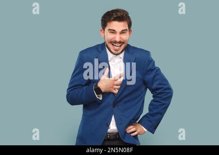 Uomo ridendo estremamente felice con baffi in piedi tenendo la mano sul petto, ascoltando scherzo divertente, guardando la macchina fotografica, indossando camicia bianca e giacca. Studio in interni isolato su sfondo azzurro. Foto Stock