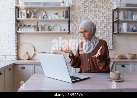 Donna in hijab a casa sconvolto e deluso leggere il messaggio dallo schermo del notebook, donna musulmana ha ricevuto cattive notizie on-line dal computer che lavora in cucina, donna araba scioccata e preoccupata triste Foto Stock