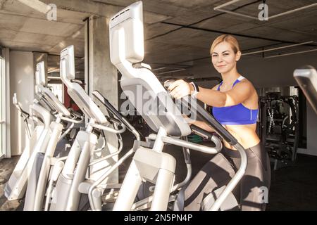 Ritratto di giovane donna atletica in forma utilizzando ellittica trainer in un centro fitness, guardando la macchina fotografica e sorridendo, indossando top sportivo e collant. Ripresa in interni con finestra sullo sfondo. Foto Stock