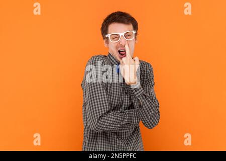 Ritratto dell'uomo nerd mettere il dito nel naso, ingannare intorno, cattive abitudini, comportamento irrispettoso, indossa una camicia con cravatta blu e occhiali bianchi. Studio al coperto isolato su sfondo arancione Foto Stock