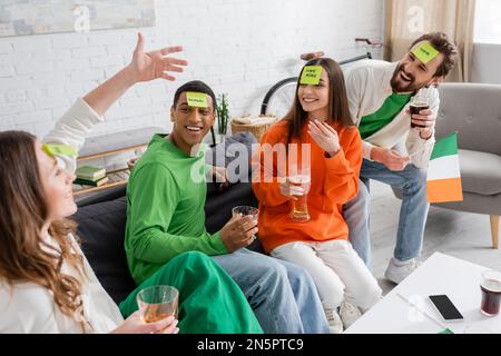 Amici interrazziali felici con note appiccicose sulla fronte guardando la donna mentre giocando indovinate chi gioco il giorno di San Patrizio Foto Stock