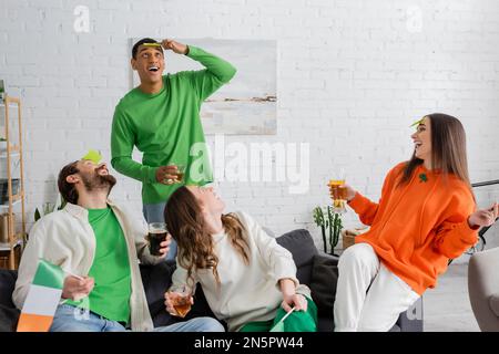 Amici felici che guardano l'uomo afroamericano che guarda la nota appiccicosa sulla fronte mentre giocano indovinate chi gioca il giorno di San Patrizio Foto Stock