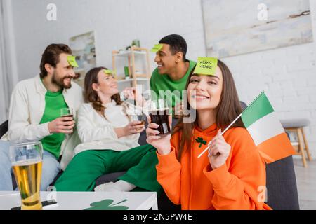 Donna felice con la parola del re sul biglietto appiccicoso che tiene la bandiera irlandese e un bicchiere di birra vicino agli amici interracial il giorno di San Patrizio Foto Stock