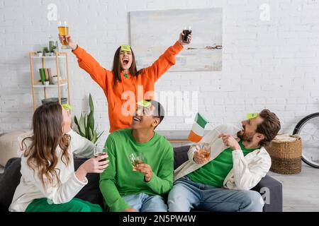 Donna eccitata che tiene la birra vicino agli amici interracial con la bandiera irlandese che gioca chi sono nel paese Foto Stock