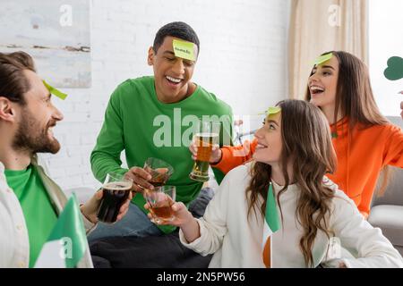 Amici multietnici felici con note appiccicose sulla fronte che si aggraffano bevande alcoliche e giocare indovinate chi partita il giorno di San Patrizio Foto Stock