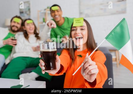 Donna stupita con nota appiccicosa sulla fronte con bandiera irlandese e bicchiere di birra vicino agli amici interracial il giorno di San Patrizio Foto Stock