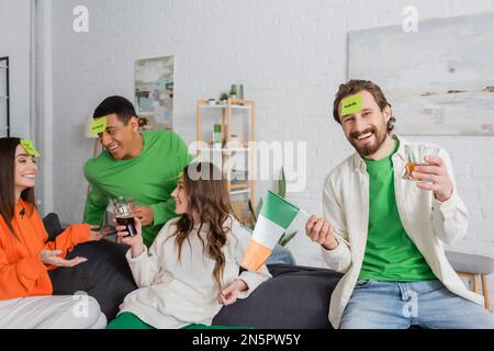 Felice uomo bearded con la parola della mela sul biglietto appiccicoso che tiene la bandiera irlandese e bere vicino agli amici interrazziali il giorno di San Patrizio Foto Stock