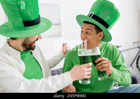 Amici interrazziali eccitati che si aggirano sulla birra verde durante il giorno di san patrizio a casa Foto Stock