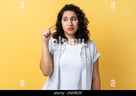 Piccola quantità. Ritratto di donna con capelli ondulati scuri che mostrano un piccolo gesto con la mano e guardando pleadingly, faccia accigliata. Studio al coperto isolato su sfondo giallo. Foto Stock