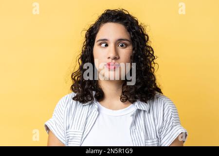 Ritratto di donna divertente con i capelli ondulati scuri che fanno il volto sciocco comico con gli occhi incrociati, pensando intensamente cercando muto e confuso. Studio al coperto isolato su sfondo giallo. Foto Stock