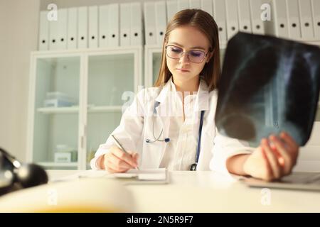 Il giovane medico in occhiali controlla le scansioni radiografiche e prende appunti nei documenti medici. Foto Stock