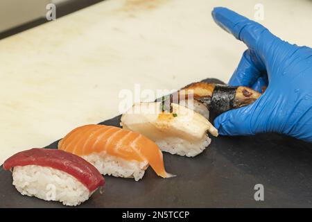 Le mani dei guanti di un cuoco che mette il nigiris appena fatto su un vassoio nero dell'ardesia Foto Stock