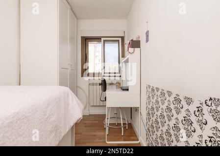 Camera da letto stretta con un letto singolo, una scrivania bianca, un armadio con porte bianche e una finestra in alluminio Foto Stock