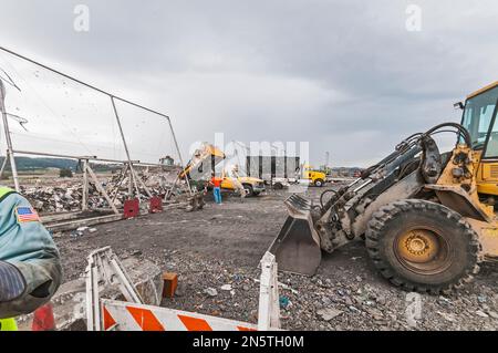 Una manica per l'operatore, una pala cingolata in primo piano, con un dumper che scarica in background mentre un operatore guarda una discarica attiva. Foto Stock