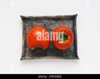 Frutta di persimmon su piatto di ceramica blu su sfondo bianco, vista da primo piano del tavolo. Immagine dettagliata di due frutti kaki con foglia, direttamente sopra la foto. Foto Stock