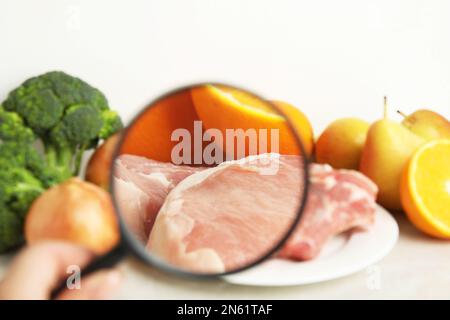 Donna con lente d'ingrandimento esplorando carne cruda e frutta, primo piano. Rilevamento di veleni Foto Stock