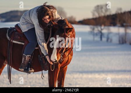 Ritratto di una donna equestre che cavalca a cavallo di fronte a un paesaggio rurale innevato d'inverno durante il tramonto all'aperto Foto Stock