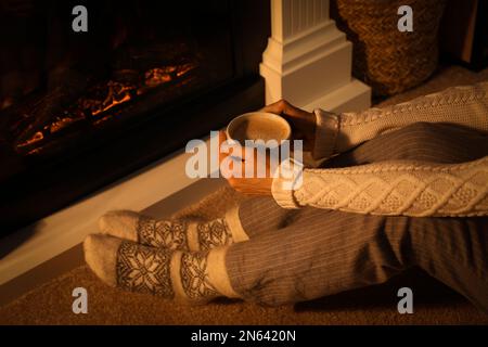 Donna in calze calde tenere bevanda calda vicino al camino con legna in fiamme a casa, primo piano Foto Stock