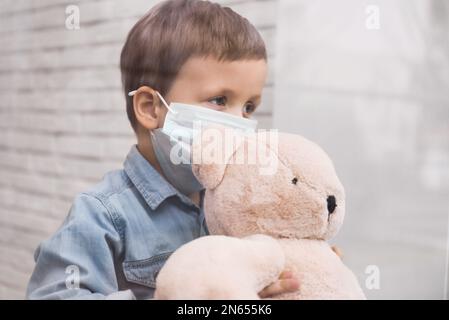 Triste ragazzino in maschera protettiva che guarda fuori dalla finestra interna, vista dall'esterno. Rimanere a casa durante la pandemia di coronavirus Foto Stock