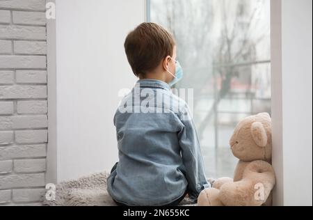 Bambino triste in maschera protettiva guardando fuori dalla finestra all'interno. Rimanere a casa durante la pandemia di coronavirus Foto Stock