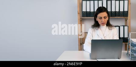 Medico di sesso femminile che lavora con il suo computer portatile in un ufficio medico seduto al tavolo. Preparazione delle informazioni dei pazienti per l'incontro con l'équipe medica in ospedale. Foto Stock