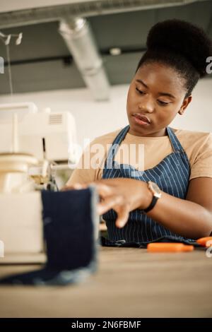 Tessuto da cucire afro-americano su misura. Giovane tessuto jeans con cuciture di design. Stilista di moda che usa una macchina da cucire nel suo studio. Creativo Foto Stock