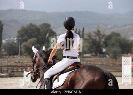 Una giovane ragazza snella rider è impegnata in equitazione. Ippodromo all'aperto con vista sulle montagne. Bellissimo cavallo di razza purosangue. Foto Stock