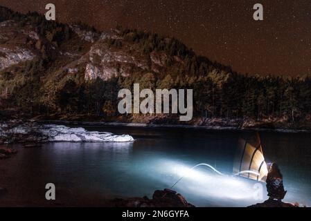 La silhouette di una ragazza di pescatori con una lanterna ondeggia di notte una canna da pesca sulle rive del fiume Katun sotto una montagna con stelle ad Altai. Foto Stock