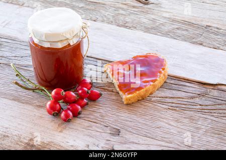 Marmellata fatta in casa di rosa canina in vaso e fetta di pane con spalmatura su tavola di legno con frutta matura Foto Stock