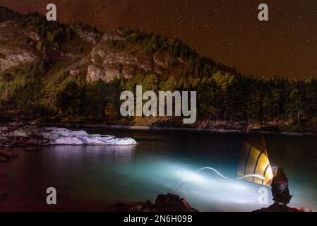 La silhouette di una ragazza di pescatori con una lanterna ondeggia di notte una canna da pesca sulle rive del fiume Katun sotto una montagna con stelle ad Altai Foto Stock