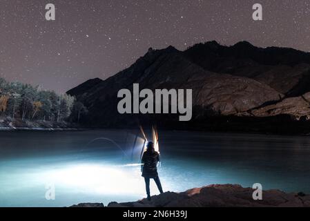 La silhouette di una ragazza pescatore con una lanterna ondeggia una canna da pesca di notte sulla riva del fiume sotto una roccia con stelle in Altai Foto Stock