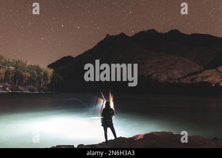 La silhouette di una ragazza pescatore con una lanterna ondeggia una canna da pesca di notte sulla riva del fiume sotto una roccia con stelle in Altai in Siberia. Foto Stock