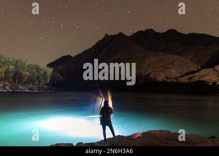 La silhouette di una ragazza pescatore con una lanterna di luce ondeggia una canna da pesca di notte sulla riva del fiume sotto una roccia con stelle in Altai in Siberia. Foto Stock