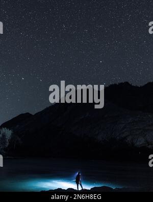 Silhouette di una ragazza pescatore con la luce di una lanterna di notte con una canna da pesca sulle rive del fiume Katun sotto una montagna con una stella Foto Stock