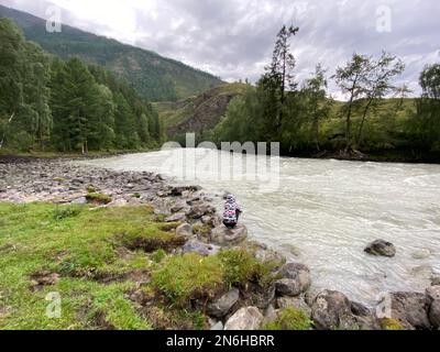 Una ragazza pescatore siede su una pietra vicino ad un fiume veloce di montagna durante il giorno. Foto Stock