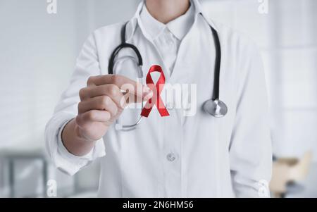 Medico tenendo nastro di consapevolezza rosso su sfondo sfocato, primo piano. Giornata mondiale della malattia dell'AIDS Foto Stock