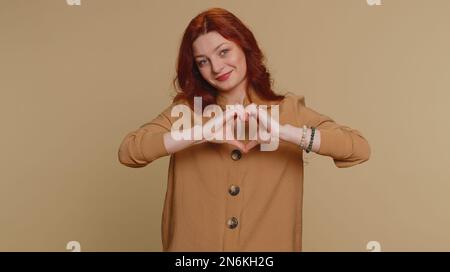 Donna innamorata. Sorridente donna rossa 20s in blusa fa gesto cardiaco dimostra segno d'amore esprime buoni sentimenti e simpatia. Giovane bella ragazza adulta isolata da sola su sfondo beige studio Foto Stock