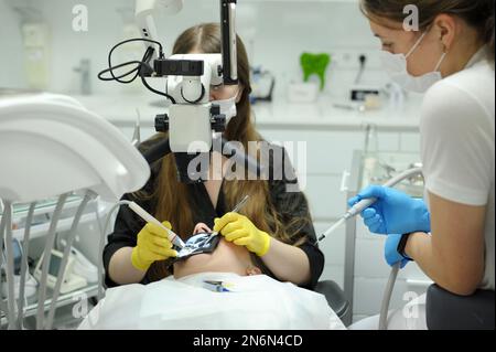 Attrezzatura odontoiatrica moderna tecnica di applicazione di guanti da dentista femmina con microscopio dentale per esaminare i denti dei pazienti medico paziente assistente dentistico tr Foto Stock