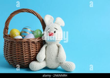 Coniglio giocattolo in maschera di protezione vicino al cesto di vimini con uova su sfondo azzurro, spazio per il testo. Vacanze di Pasqua durante la quarantena COVID-19 Foto Stock