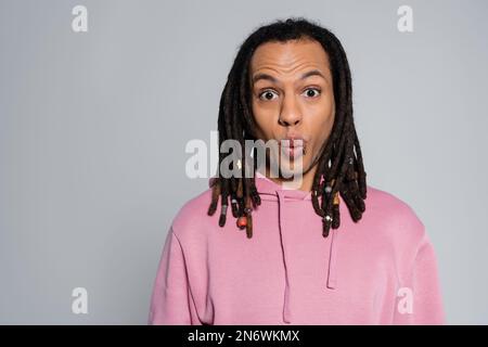 scioccato uomo multirazziale in rosa con cappuccio che perlustra le labbra e guarda la fotocamera isolato sul grigio Foto Stock