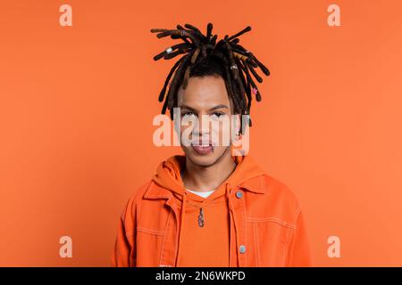 ritratto di uomo multirazziale trafitto con dreadlocks guardando la macchina fotografica isolata su sfondo corallo Foto Stock