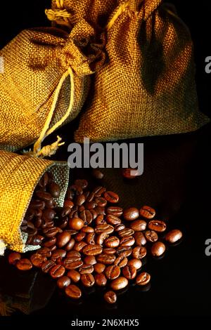 Sacchi di Hessian pieni di chicchi di caffè tostati che fuoriescono da un sacco aperto sul vetro nero Foto Stock