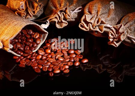 Sacchi di Hessian pieni di chicchi di caffè tostati che fuoriescono da un sacco aperto sul vetro nero Foto Stock