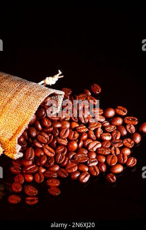 Sacchi di Hessian pieni di chicchi di caffè tostati che fuoriescono da un sacco aperto sul vetro nero Foto Stock