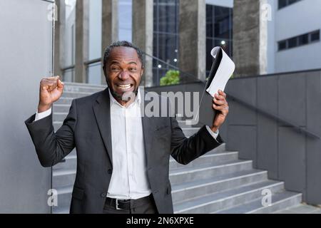 Felice uomo d'affari afro-americano, avvocato, banchiere in piedi vicino all'ufficio e in possesso di una cartella con documenti, ha ricevuto buone notizie. Allegro sorride alla telecamera, alza le mani in vittoria. Foto Stock